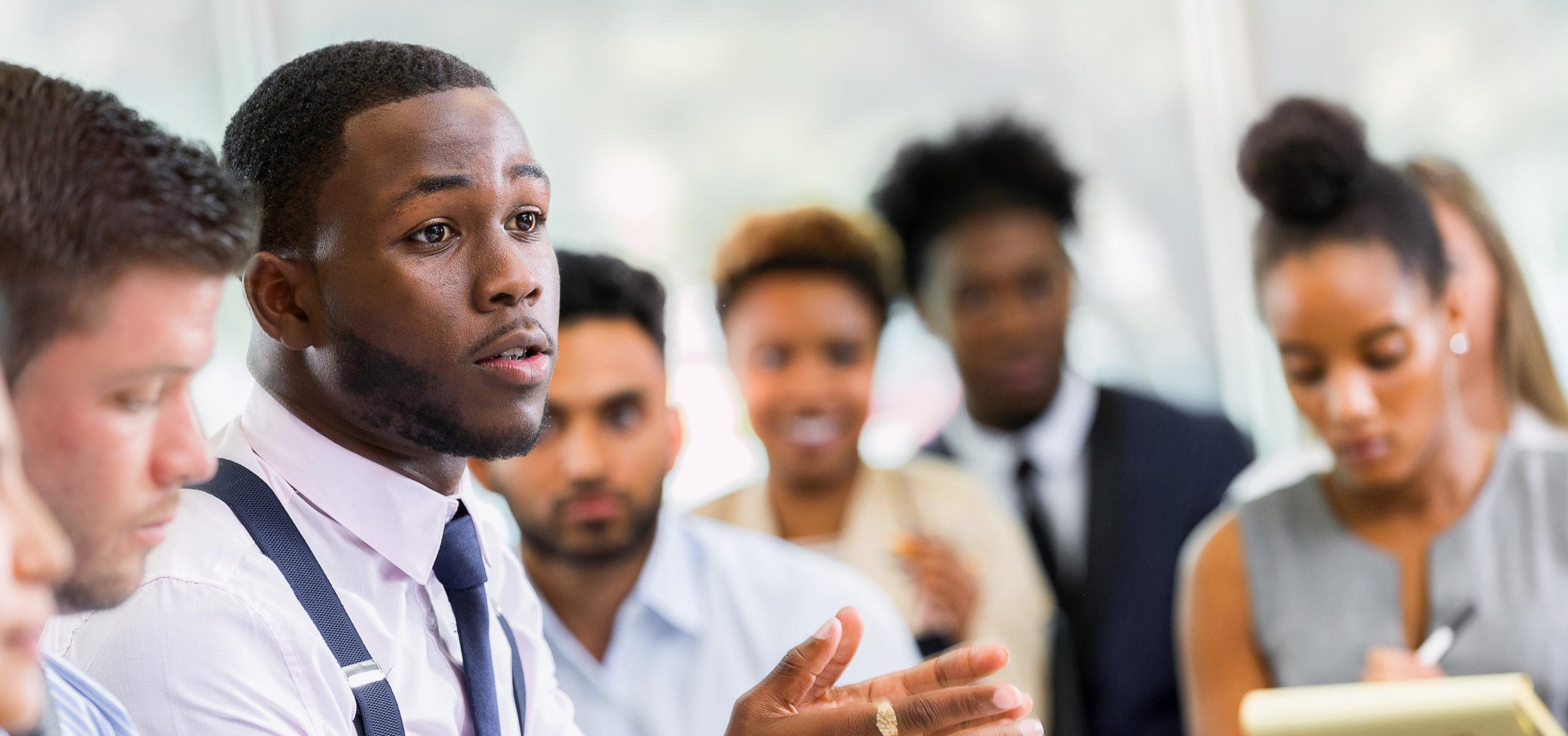 A serious young African American male business student talks with his hands as he sits at a table in a lecture hall and poses a question to an unseen professor. Other students sit in the audience in the background.