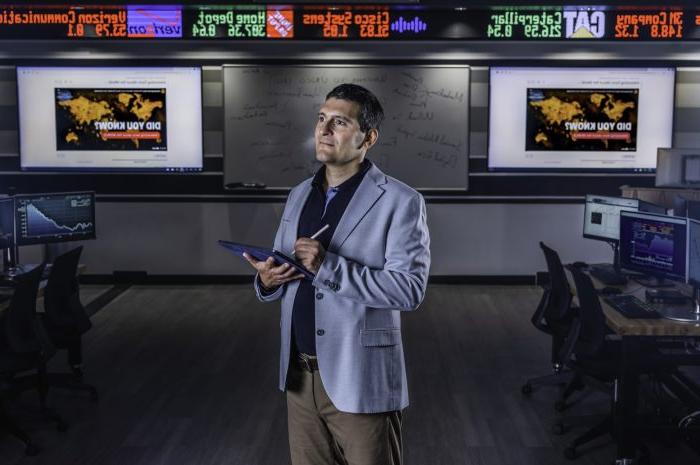 A man in a blazer stands in a modern office with multiple computer monitors and flat screens behind him displaying a world map and the text "Did You Know?". He holds a tablet and looks thoughtful. An electronic stock ticker with various company names and indices runs above.