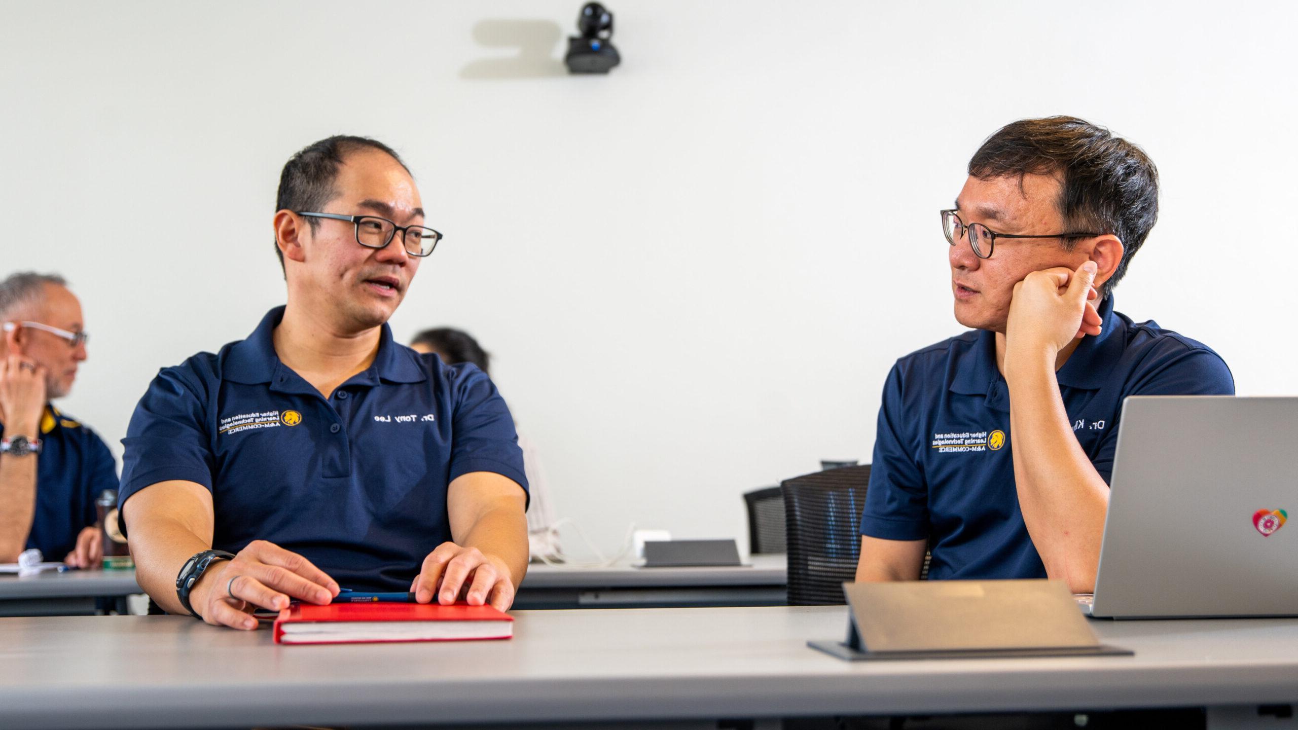 Two men in navy blue polo shirts sit at a table in a meeting. One listens with his hand on his face, while the other, holding a red notebook, speaks. A laptop is in front of them.