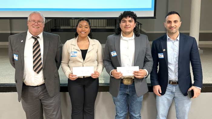 Four people stand shoulder to shoulder for a photo opportunity. The two people in the middle are students holding their scholarship envelopes.